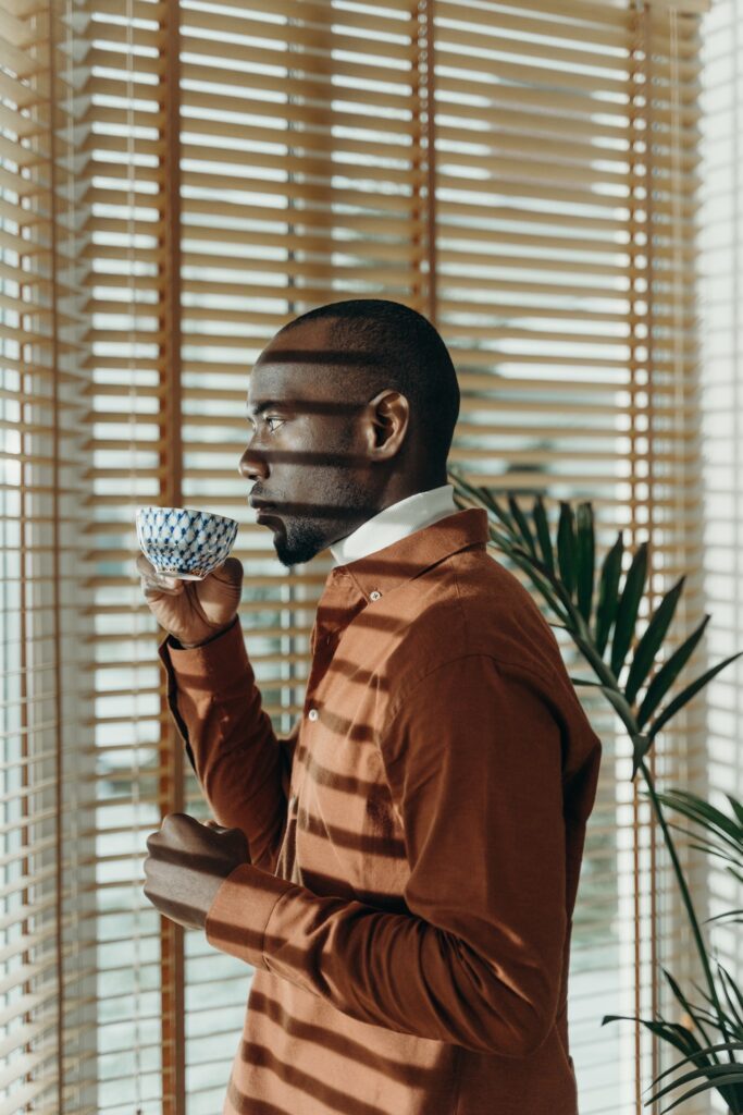 Hombre de piel oscura con cafe en mano viendo a traves de su ventana